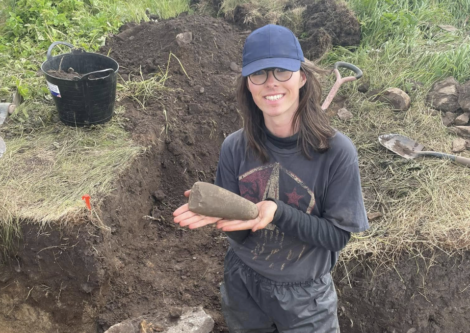 A person wearing glasses and a cap stands at an excavation site, holding a large, elongated stone artifact with both hands. Soil, tools, and a bucket are visible nearby.