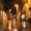 A wet, empty street at night with string lights overhead and shop windows illuminated on both sides.