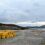 A paved industrial area near a body of water with stacks of yellow barrels, distant hills, and an overcast sky.