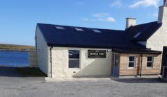 A light-colored building with a dark roof labeled "Brough Store" stands near the water under a partly cloudy sky.