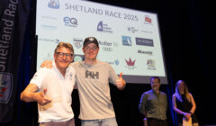 Two men pose with thumbs up in front of a Shetland Race 2025 sign, with two people standing and event sponsor logos displayed on a screen behind them.