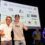 Two men pose with thumbs up in front of a Shetland Race 2025 sign, with two people standing and event sponsor logos displayed on a screen behind them.