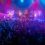 A band performs on stage under colorful lights while a large crowd watches and dances in front of the stage at an indoor concert venue.