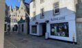 White-fronted shop with "Burns Lane Clothing" signage and a blue-and-white bunting-decorated window, on a paved street in a stone-built