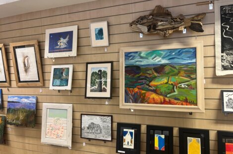 Various framed artworks, including paintings, drawings, and a wooden fish sculpture, are displayed on a light wooden wall in a gallery setting.
