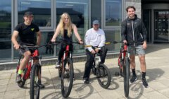 Four people are posing outside a building with their bicycles, three with mountain bikes and one with a BMX, on a paved area in front of large windows.