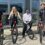 Four people are posing outside a building with their bicycles, three with mountain bikes and one with a BMX, on a paved area in front of large windows.