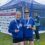 Two women in blue athletic gear smile and display medals in front of a Kilmarnock Harrier & Athletic Club tent at an outdoor event.