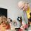 A teacher helps a student at their desk in a classroom with a chalkboard showing geometric shapes in the background.