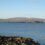 A rocky shoreline with calm water, distant low hills, and a clear blue sky.