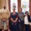 Six people stand side by side indoors in front of two large stained glass windows, posing for a group photo.