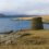 A round stone tower stands on grassy ground beside a rocky shoreline, with calm water and hills visible in the background under a cloudy sky.