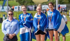 Four people stand outdoors; three women wearing medals and draped in blue and white flags, and one man in sunglasses and a blue shirt. The setting appears to be a sports event.