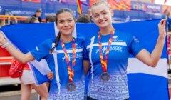 Two female athletes wearing blue shirts and medals hold a blue and white flag, smiling at an outdoor sporting event.