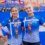 Two female athletes wearing blue shirts and medals hold a blue and white flag, smiling at an outdoor sporting event.