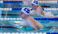 Two swimmers in a pool compete in a breaststroke race, both wearing swim caps with a blue and white flag design.