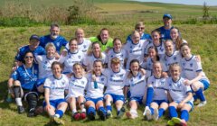 A women’s soccer team and coaches pose for a group photo on grass, wearing matching white and blue uniforms, with fields and hills in the background.
