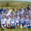 A women’s soccer team and coaches pose for a group photo on grass, wearing matching white and blue uniforms, with fields and hills in the background.