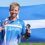 A man in a blue Team Shetland shirt holds a medal and mascot, standing in front of a large Shetland flag on a sunny day.