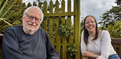 An older man and a younger woman sit smiling on a wooden bench outdoors in front of a yellow garden fence and greenery.