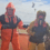 Two people in waterproof fishing gear and gloves stand on a boat deck at sea, smiling. Seagulls fly in the sky above them.