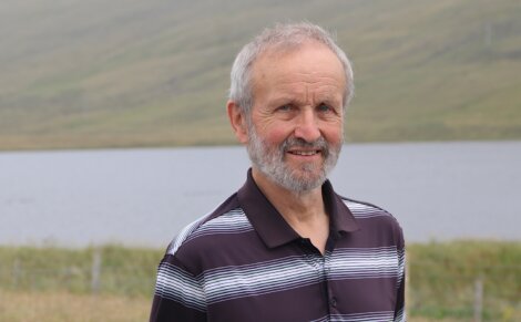An older man with a gray beard stands outdoors in front of a lake and grassy hills, wearing a black and white striped polo shirt.