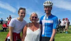 Three people pose outdoors on grass; one wears cycling gear and helmet, the others wear casual clothes. Other people, bikes, and a blue sky are visible in the background.