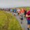 A large group of people of various ages running outdoors on a paved path beside a grassy field under an overcast sky.