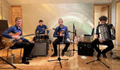 Four young musicians sit with their instruments—electric guitar, drum set, violin, and accordion—in a simple indoor performance space with microphones in front of them.