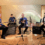 Four young musicians sit with their instruments—electric guitar, drum set, violin, and accordion—in a simple indoor performance space with microphones in front of them.