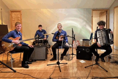 Four young musicians sit with their instruments—electric guitar, drum set, violin, and accordion—in a simple indoor performance space with microphones in front of them.