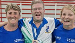 Three people stand smiling in front of a sign for the Scotland Disability Sport National Learning Games. The man in the center wears a medal and a blue shirt; the two women wear matching blue shirts.