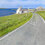 A narrow road marked A970 curves beside the sea, bordered by grassy slopes, with a few stone buildings near the shoreline under a partly cloudy sky.