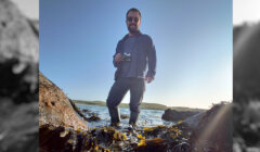 A person wearing sunglasses and holding a camera stands in shallow water between rocks and seaweed, with a clear blue sky and distant hills in the background.
