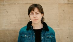 A person with short brown hair wearing a denim jacket stands in front of a beige stone wall, looking at the camera with a neutral expression.