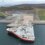 A large red and white ship with a helipad is docked at a remote, industrial pier surrounded by hilly landscape and water under an overcast sky.