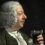 A man with white curly hair and a green coat holds a glass of dark liquid, looking at it with a slight smile.
