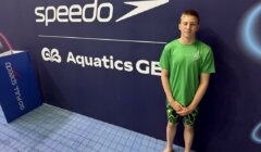 A young swimmer in a green shirt and swim trunks stands by a wall with "Speedo" and "Aquatics GB" logos at an indoor poolside.