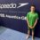 A young swimmer in a green shirt and swim trunks stands by a wall with "Speedo" and "Aquatics GB" logos at an indoor poolside.