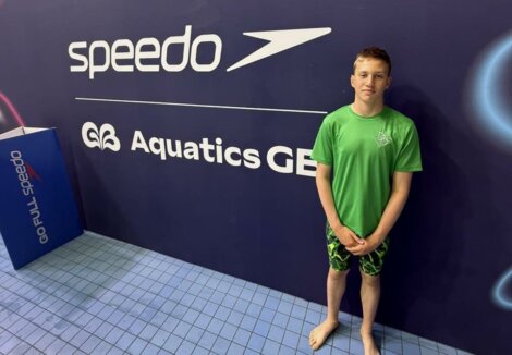 A young swimmer in a green shirt and swim trunks stands by a wall with "Speedo" and "Aquatics GB" logos at an indoor poolside.