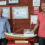 Two men stand beside a detailed model boat displayed indoors, with informational signs and framed photos on the red wall behind them.