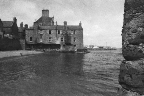 Black and white photo of a stone building by the water, with chimneys on the roof, a small beach, and a person and dog in the background.