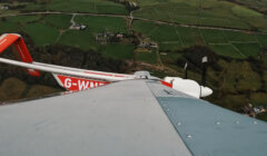 A view from an aircraft wingtip shows the tail and propeller in flight over green fields and farmland, with the "G-WAND" registration visible on the tail.