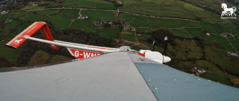 A view from an aircraft wingtip shows the tail and propeller in flight over green fields and farmland, with the "G-WAND" registration visible on the tail.