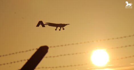 A small airplane flies at sunset above barbed wire, with the sun low on the horizon and a clear sky in the background.