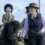 A young child in riding gear smiles while sitting on a small black pony, guided by two adults in equestrian attire during an outdoor event.