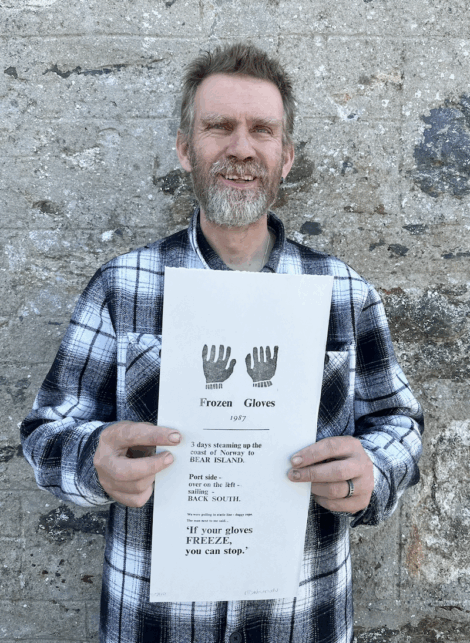 A man in a plaid shirt stands against a stone wall, smiling and holding a printed paper titled "Frozen Gloves" with glove graphics and text.