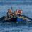 Five people are rowing together in a blue boat on open water, pulling oars in unison and displaying concentrated, determined expressions.