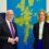 Two people in business attire stand in front of a blue and green map display mounted on a wall.