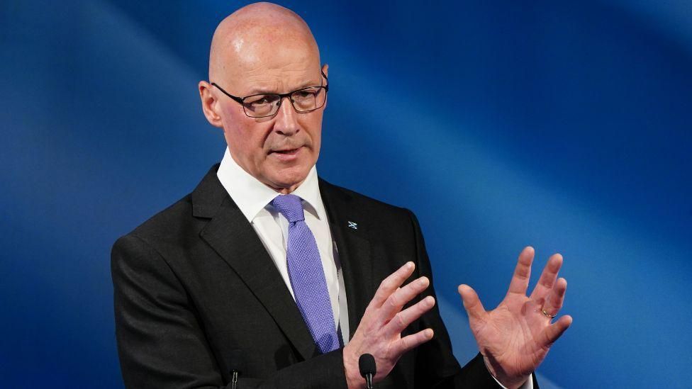 A bald man in a suit and tie gestures with his hands while speaking at a podium against a blue background.